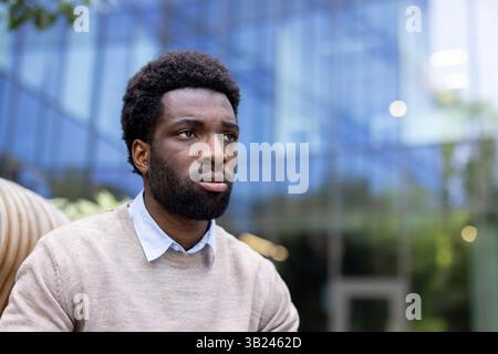 Un homme afro-américain réfléchi est capturé dans un moment de contemplation, dehors dans un cadre moderne, pensant peut-être à l'avenir. Banque D'Images
