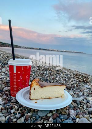 Cheesecake et tasse de café à emporter sur une plage de galets avec mer calme et ciel pastel en arrière-plan, scène de collation extérieure paisible Banque D'Images