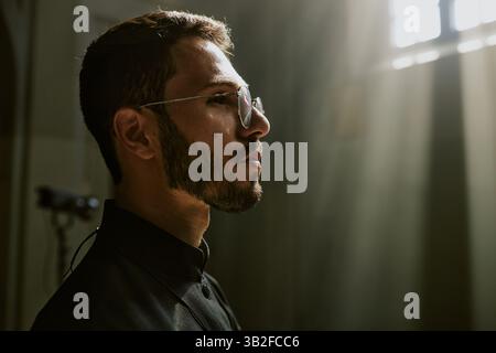 Gros plan moyen d'un jeune beau prêtre avec des lunettes rondes en robe noire regardant droit Banque D'Images