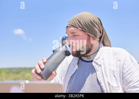 Jeune homme hispanique buvant de l'eau dans une bouteille réutilisable tout en travaillant sur son ordinateur portable à l'extérieur par une journée ensoleillée. Concept : mode de vie sain d'un numérique Banque D'Images