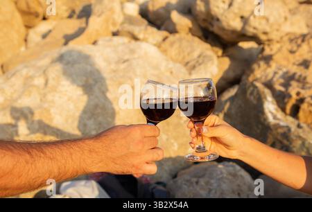 Gobelets en verre avec du vin rouge dans les mains de deux personnes sur fond d'un rocher illuminé par le soleil Banque D'Images