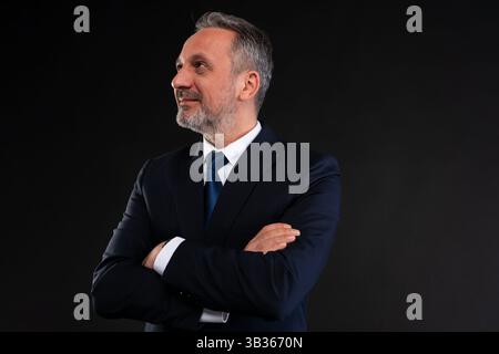 Homme d'affaires mature en costume avec les bras croisés, regardant sur le côté avec une expression confiante et réfléchie, sur fond noir. Banque D'Images