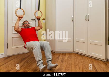 Un homme effectue un exercice d'aviron en utilisant des anneaux de gymnastique à l'intérieur. Il porte une chemise rouge et un pantalon de survêtement gris, mettant en valeur sa force et sa forme physique ROU Banque D'Images