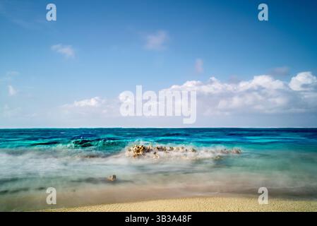 Scène côtière avec des eaux turquoises se lavant doucement sur les rochers, créant un effet brumeux. Le ciel est bleu vif avec des nuages blancs dispersés. Banque D'Images