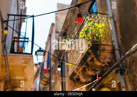 Des chaînes d'ampoules colorées, sont enfilées à travers une ruelle étroite entre de vieux bâtiments en pierre avec balcons. La lumière du soleil illumine la scène Banque D'Images