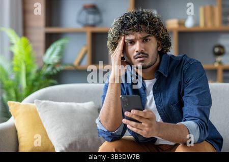 Un jeune homme inquiet regarde son téléphone tout en étant assis sur un canapé à la maison. Il tient la tête avec sa main. Banque D'Images