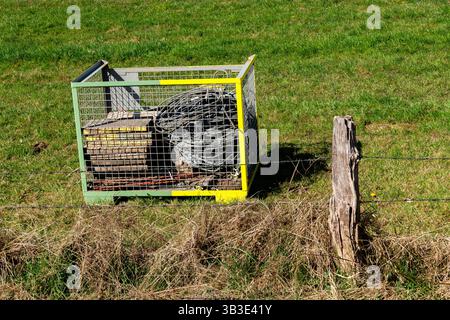 Un conteneur de stockage en métal rempli d'outils se trouve dans un champ vert éclatant sous un ciel bleu. A proximité, un poteau en bois ajoute au cadre rural, showcasin Banque D'Images