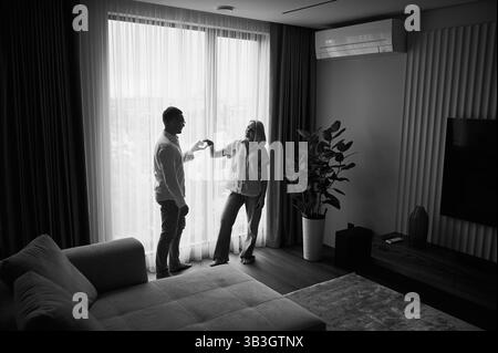 Charmant couple montrant le symbole du cœur. Jeune homme et sa petite amie debout en face de la fenêtre à la lumière naturelle à la maison. Mignons jeunes mariés heureux de leur nouvel appartement. Image en noir et blanc. Banque D'Images