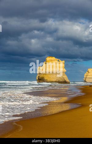 Les vagues s'écrasent sur le rivage sablonneux tandis que les majestueuses formations rocheuses des douze Apôtres s'élèvent de la mer sous un ciel spectaculaire. Banque D'Images
