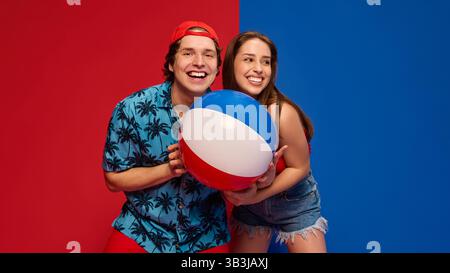 Couple joyeux, homme et femme riant et posant avec un ballon de plage, montrant des vibrations positives d'été sur fond rouge et bleu fendu Banque D'Images
