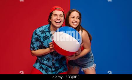 Couple joyeux, homme et femme riant et posant avec un ballon de plage, montrant des vibrations positives d'été sur fond rouge et bleu fendu Banque D'Images