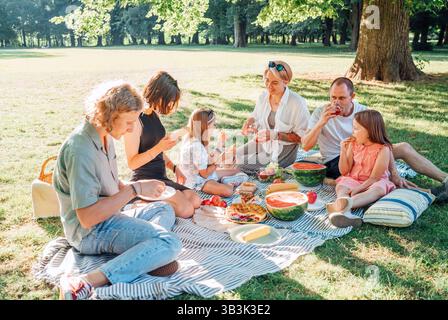 Grande famille assise sur une couverture de pique-nique sous les tilleuls dans un parc de la ville le dimanche ensoleillé. Ils bavardent et mangent du maïs bouilli et de la pastèque. Famil Banque D'Images
