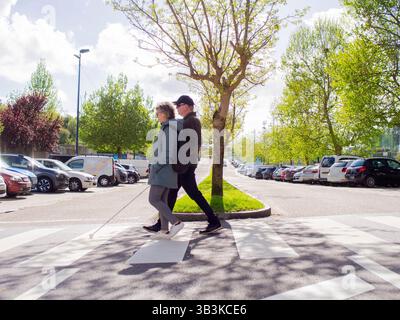 vue de côté d'un homme sans vision marchant sur la traversée du zèbre en utilisant sa canne et une femme fournissant des conseils. Banque D'Images