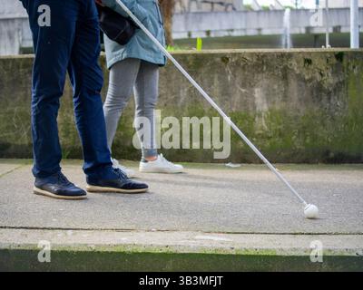 Homme aveugle marchant sur le trottoir en utilisant sa canne et une femme fournissant des conseils. Banque D'Images