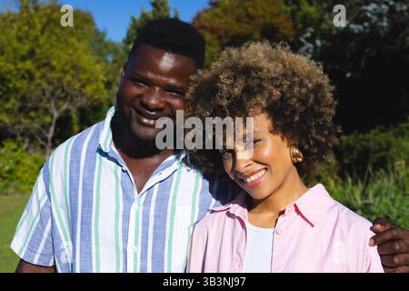 Debout couple diversifié embrassant dans le jardin ensoleillé, près des arbres feuillus et ciel bleu clair Banque D'Images