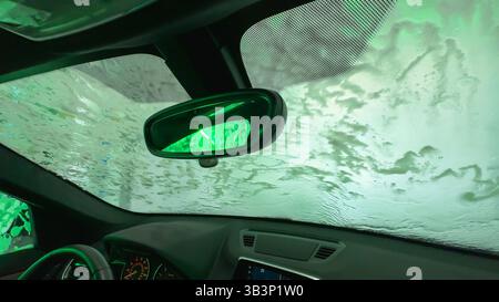 Vue intérieure d'une voiture à l'intérieur d'un lave-auto avec des lumières vertes et de l'eau savonneuse sur le pare-brise. Banque D'Images