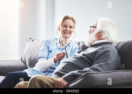 Médecin mûr aidant l'homme âgé à la maison avec un stéthoscope. Professionnelle de santé féminine examinant la fréquence cardiaque d'un vieux patient masculin sur un canapé. Femme Banque D'Images