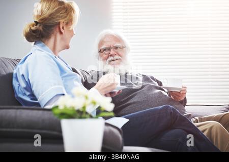 Homme senior à la retraite et infirmière se relaxant, parlant et se liant sur le canapé dans une maison de vieillesse tout en buvant du café. Aimable soignant et homme apprécient Banque D'Images