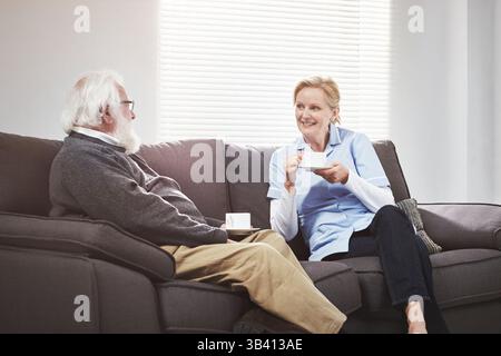 Heureux soignant et son patient senior buvant du café et ayant une conversation agréable et chaleureuse sur un canapé. Une infirmière à domicile aide à la liaison et Banque D'Images