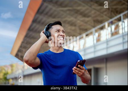 Un homme asiatique en t-shirt bleu porte des écouteurs et écoute de la musique tout en tenant son téléphone dans un stade de sport. Détente en plein air. Exercice ou baguette Banque D'Images