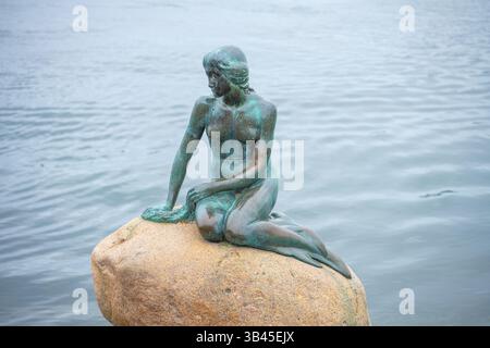 Une statue en bronze d'une sirène se trouve gracieusement sur un rocher au bord de l'eau à Copenhague. Les visiteurs admirent la sculpture emblématique, symbole de la ville et inspirée par un conte. Banque D'Images