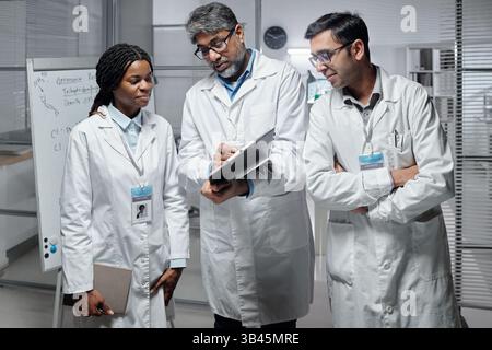 Groupe de trois professionnels de la santé divers discutant de l'information du patient tout en se tenant debout dans un bureau moderne avec des fournitures de bureau sur la table et le tableau blanc en arrière-plan Banque D'Images