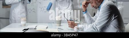Médecin âgé assis à une table blanche avec des livres ouverts, tenant des lunettes et regardant vers le bas. Autres professionnels de la santé en arrière-plan discutant Banque D'Images