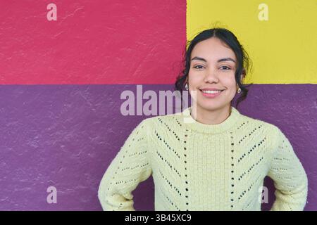 Portrait d'une jeune femme latine joyeuse portant un pull vert clair, posant contre un mur vibrant, peint géométriquement avec violet, rouge, an Banque D'Images