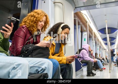 Deux jeunes femmes sont assises dans un train de métro, utilisent leur smartphone et écoutent de la musique avec des écouteurs pendant leur trajet quotidien Banque D'Images