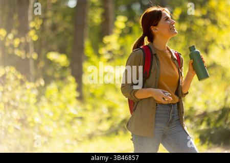Une jeune femme marche à travers une forêt verdoyante, tenant une bouteille d'eau et souriant tandis que la lumière du soleil filtre à travers les arbres. Banque D'Images