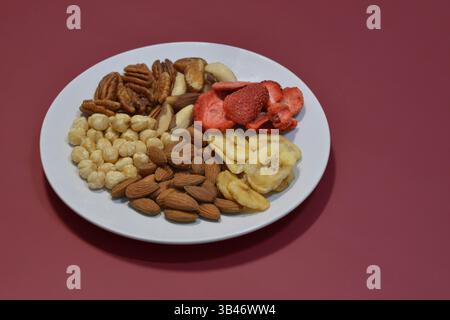 Une variété de noix et de fruits secs sur une assiette sur fond rouge. Noisettes, noix de pécan, noix du brésil, amandes, fraises séchées et bananes. Le concept Banque D'Images