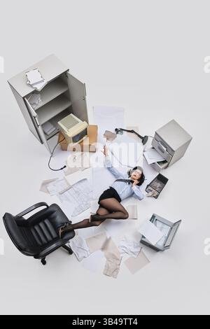 Photo vue aérienne. Employé de bureau féminin couché sur le sol avec une cravate lâche et des papiers dispersés autour dans la scène de travail chaotique. Banque D'Images