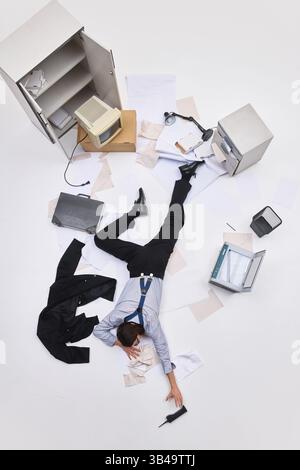 Homme d'affaires épuisé couché face vers le bas sur le sol recouvert de papier atteignant le téléphone dans la scène de bureau chaotique. Photo vue aérienne. Banque D'Images