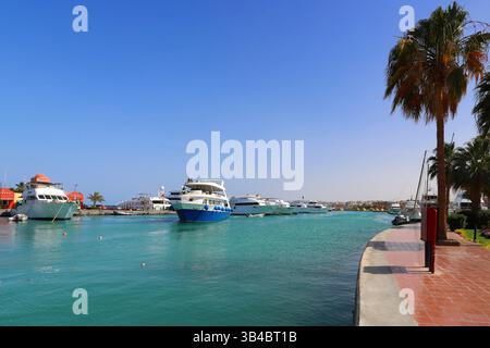 Yachts de luxe dans la marina à Hurghada, en Égypte, avec minarets mosquée et promenade bordée de palmiers sous un ciel bleu clair, scène de station balnéaire de la mer Rouge. Banque D'Images