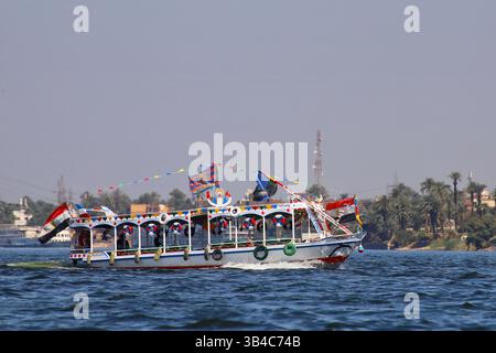 Bateau à moteur décoré traditionnel naviguant sur le Nil en Égypte avec des drapeaux égyptiens et des banderoles colorées, des palmiers en arrière-plan. Banque D'Images