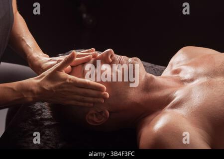 Thérapeute donnant un massage facial à un client masculin détendu dans un cadre de spa, se concentrer sur la pression douce sur le front. Banque D'Images