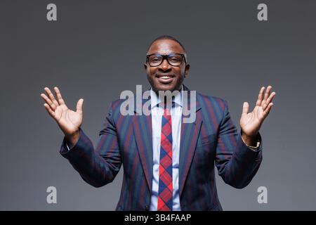 Homme d'affaires joyeux en costume rayé et lunettes, souriant avec les mains levées dans un geste hausseur d'épaule. Concept de succès, confiance, attitude positive, et Banque D'Images