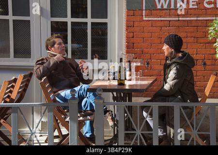 Deux personnes s'assoient à une table de café en plein air par une journée ensoleillée. Un homme dans une veste marron et un Jean est assis en face d'une femme dans une parka sombre. Bouteilles et verres Banque D'Images