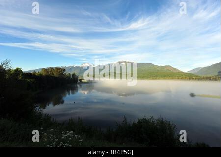 La brume matinale dérive au-dessus d'un lac de montagne immobile, reflétant le ciel bleu et les sommets enneigés dans une scène tranquille du début de l'été. Banque D'Images