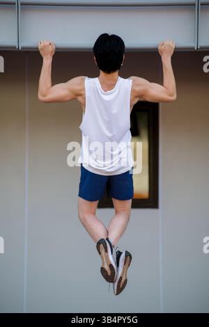 Corps complet d'un homme asiatique dans le débardeur blanc faisant un pull up sur la barre horizontale dans le stade de sport. Musculation, exercice extérieur ou entraînement. Banque D'Images