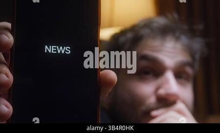 Jeune homme excité pointant vers l'écran de smartphone affichant le mot NOUVELLES, réagissant avec enthousiasme aux derniers titres dans un cadre intérieur confortable avec Banque D'Images