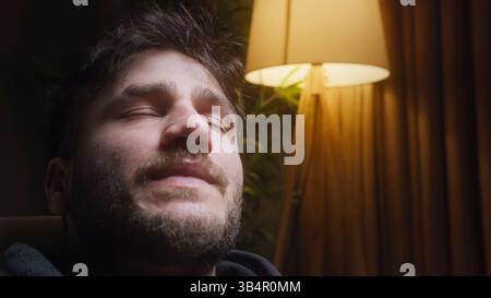 Jeune homme méditant dans une pièce faiblement éclairée avec un lampadaire chaud en arrière-plan, reflétant une ambiance calme et introspective Banque D'Images