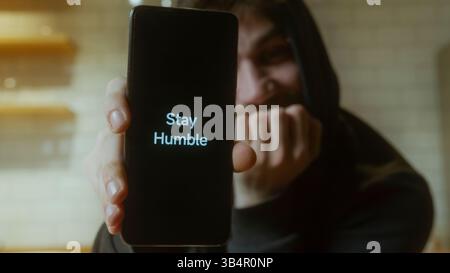 Jeune homme tenant un smartphone affichant une citation motivante restez humble dans un cadre intérieur confortable Banque D'Images