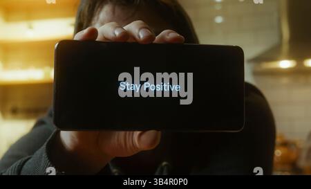 Homme souriant tenant un smartphone affichant un message Stay positive dans l'éclairage intérieur chaud Banque D'Images