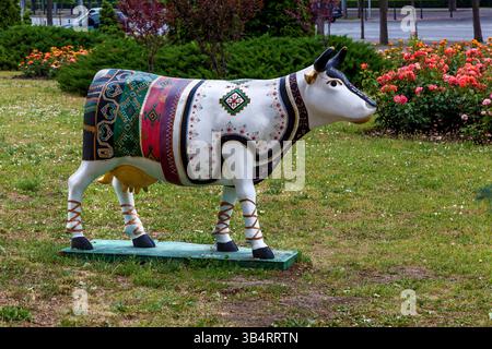 Sculpture artistique de vache peinte avec des motifs ethniques vibrants debout sur l'herbe verte près de fleurs en fleurs dans un parc de la ville Banque D'Images