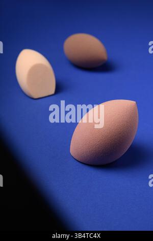 Trois mélangeurs cosmétiques différents sur un fond bleu avec une ombre contrastée. Éponges de maquillage marron avec découpes pointues. Placer pour le texte. Banque D'Images