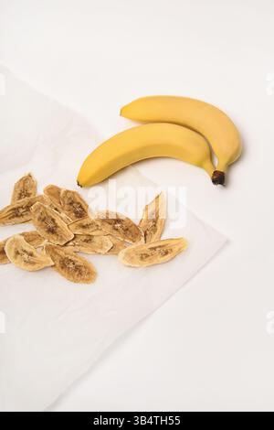 Fond blanc. Deux bananes fraîches, à côté desquelles sont séchées des tranches de bananes sur du papier parchemin. Banque D'Images