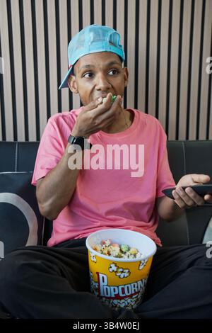 Jeune homme assis sur un canapé, mangeant du pop-corn dans un seau et tenant la télécommande, profitant du divertissement à domicile Banque D'Images