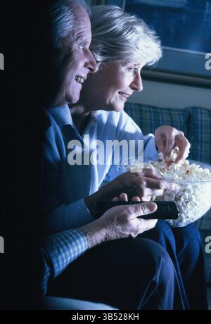Homme senior et femme mangeant du pop-corn à l'intérieur et regardant la télévision. Banque D'Images