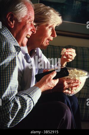 Homme senior et femme mangeant du pop-corn à l'intérieur et regardant la télévision. Banque D'Images
